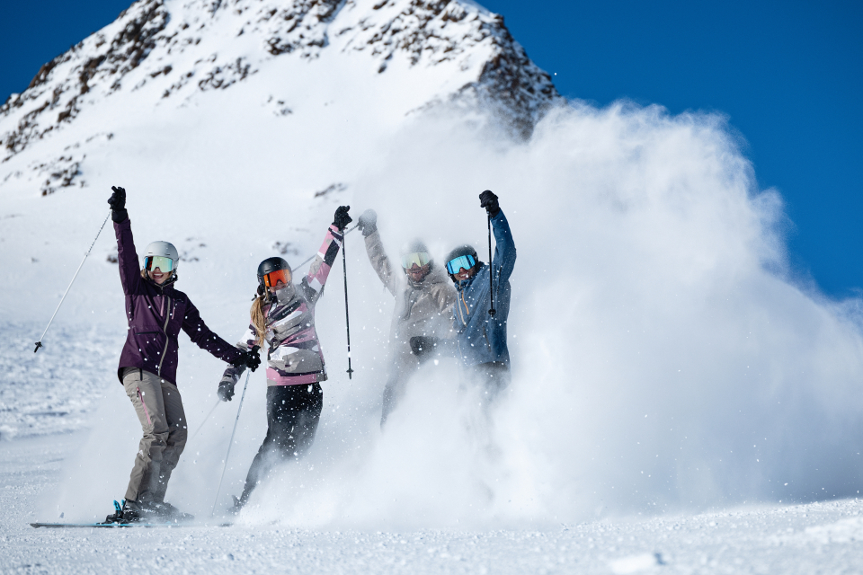 4 Skifahrer stehen in einer Schneewolke, lachen und heben die Hände