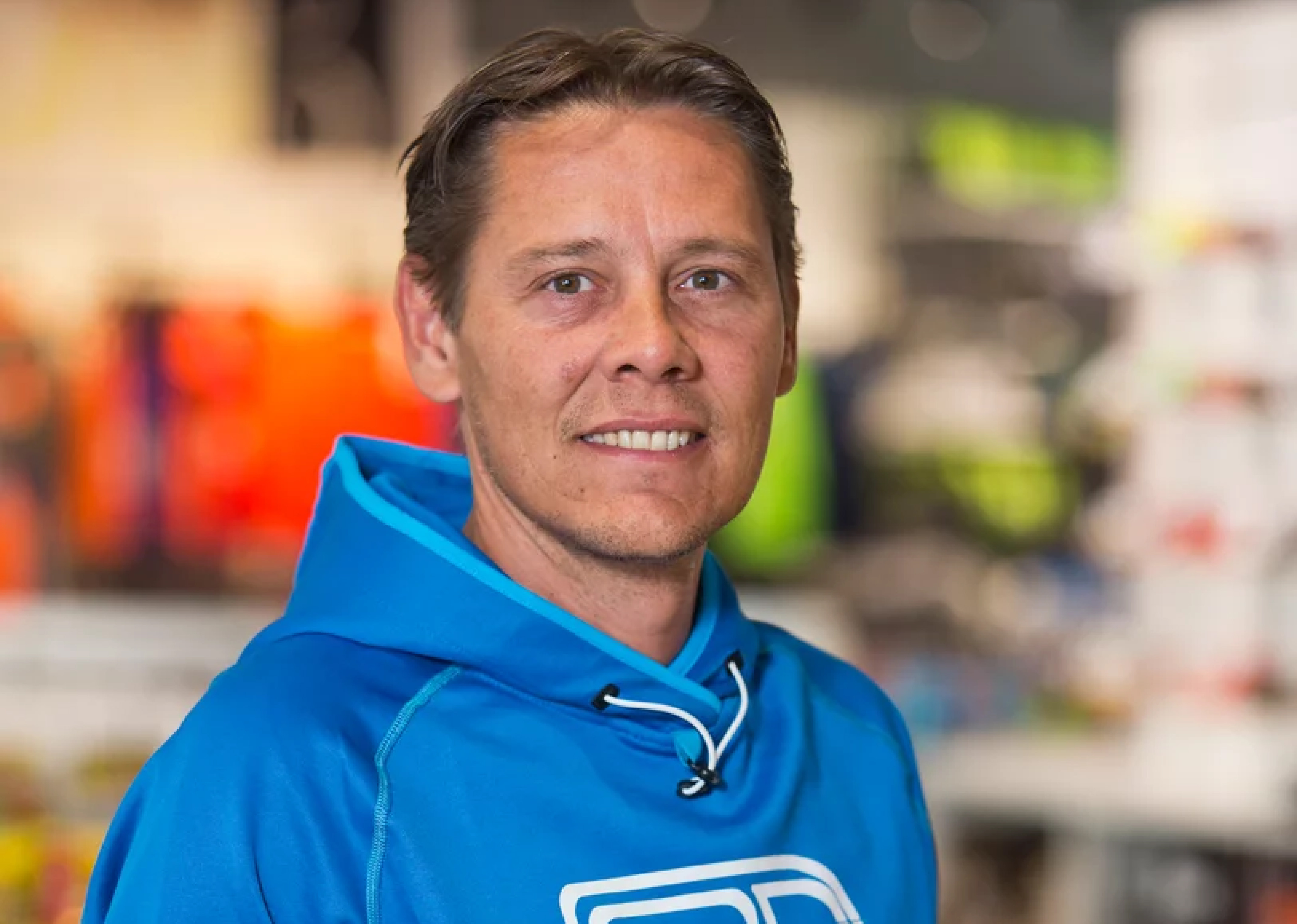 Eine Person in einem blauen Kapuzenpulli lächelt in einem Geschäft mit verschwommenen bunten Gegenständen im Hintergrund in die Kamera und vermittelt eine ungezwungene und freundliche Stimmung.