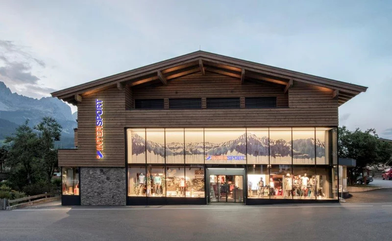 Holzgebäude im alpinen Stil mit großen Glasfenstern, verziert mit Grafiken der Berglandschaft. Auf dem Ladenschild steht „InterSport“. Der Himmel darüber ist bewölkt.