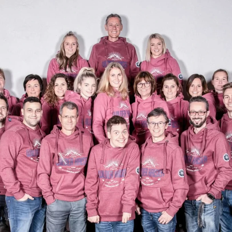 Eine Gruppe von Menschen in einheitlichen kastanienbraunen Kapuzenpullis steht zusammen und lächelt in einem gut beleuchteten Studio. Die Stimmung ist freundlich und kooperativ.
