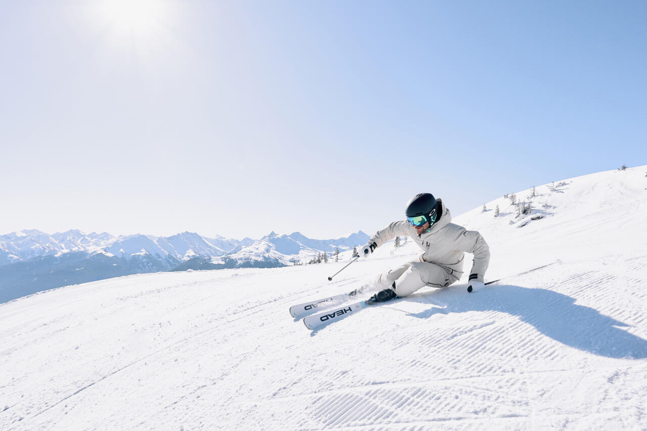 A skier wearing white clothing and a black helmet skis down the slope in sunshine and under a bright blue sky.