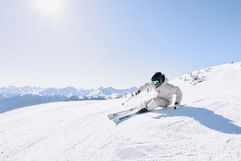 Ein Skifahrer in weißer Kleidung und schwarzem Helm fährt bei Sonnenschein und strahlend blauem Himmel die Piste hinunter