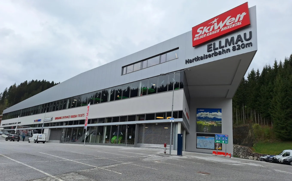 Modernes Skigebietsgebäude mit der Aufschrift „SkiWelt Ellmau” vor einem bewölkten Himmel. Hohe Kiefern säumen die rechte Seite und schaffen eine ruhige und einladende Atmosphäre.