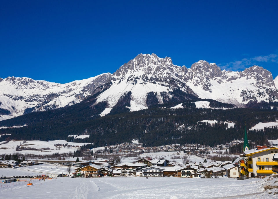 Snow-covered mountains during the day with trees on the slopes and small yellow houses and hotels in the valley below.