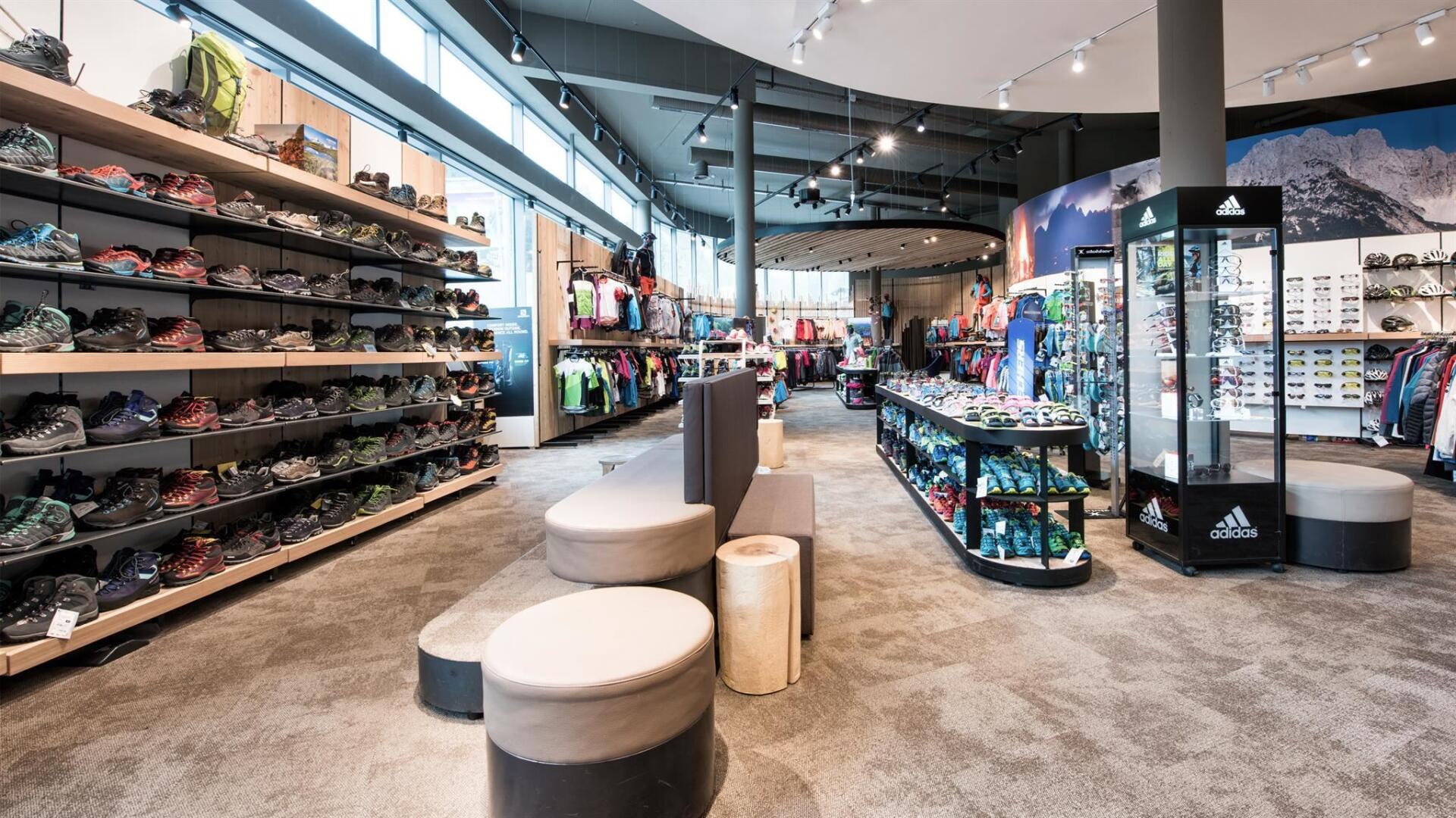 Ein hell erleuchteter Outdoor-Sportladen mit Schuhregalen auf der linken Seite, Kleiderständern mit farbenfroher Kleidung in der Mitte und Auslagen mit Sportausrüstung.