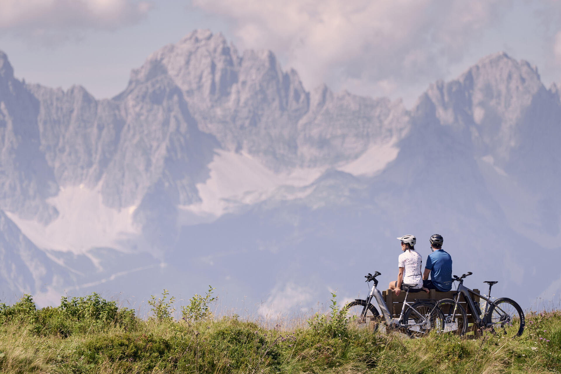 Two cyclists sit on a bench and gaze at the mountain scenery.
