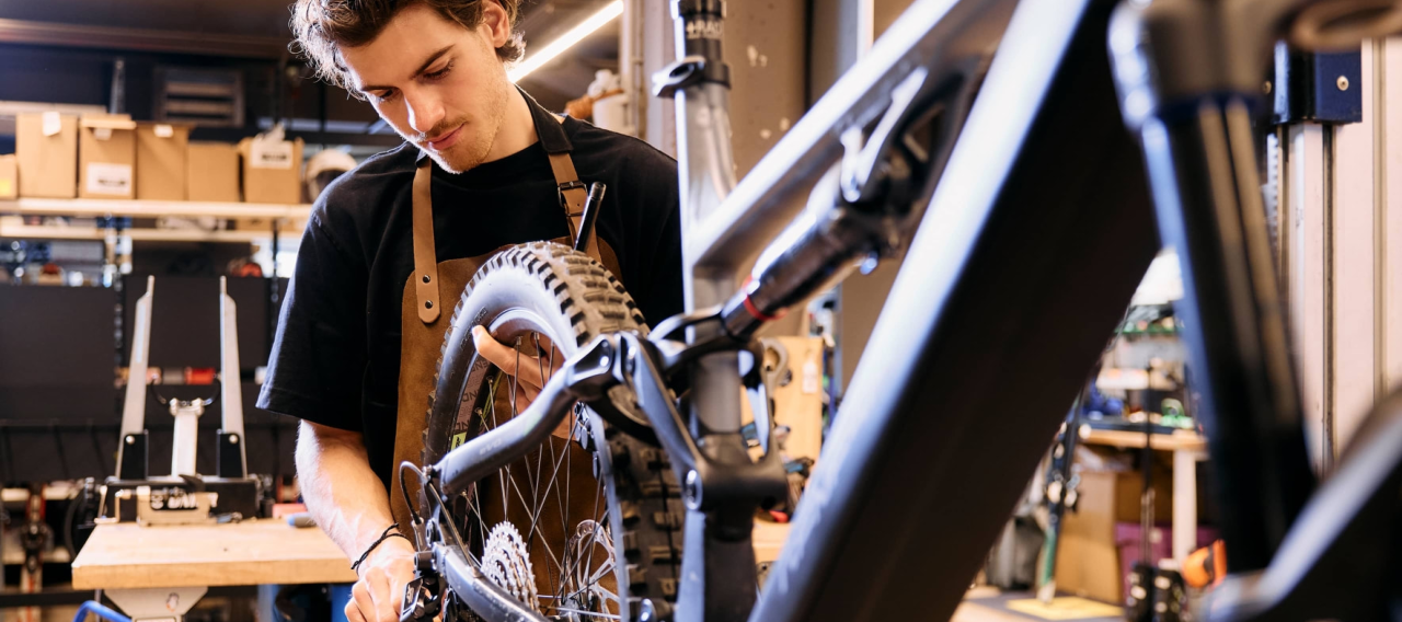 Ein junger Mechaniker in einer Werkstatt ist konzentriert damit beschäftigt, ein Fahrradrad zu reparieren. Er trägt ein schwarzes Hemd und eine braune Schürze, ist von Werkzeugen und Kisten umgeben und strahlt Konzentration aus.