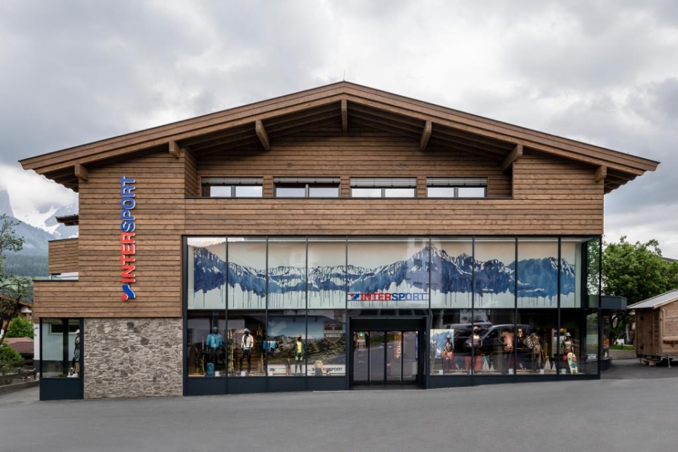 Wooden building in Alpine style with large glass windows, decorated with graphics of the mountain landscape. The shop sign reads “InterSport.” The sky above is cloudy.