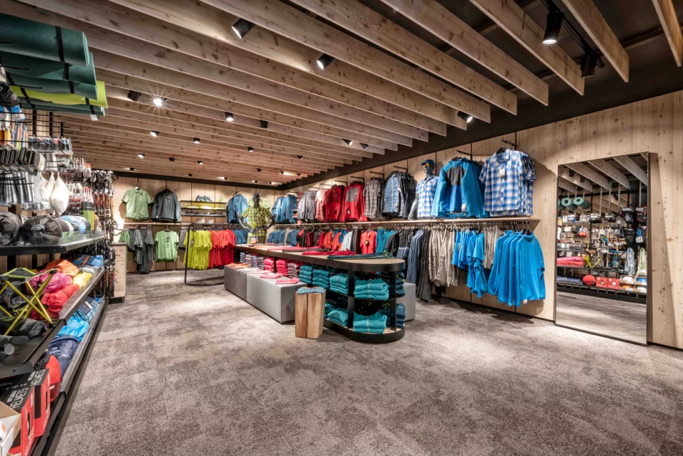 A well-lit outdoor clothing store with wooden beams on the ceiling, where colourful jackets, shirts and folded garments are neatly displayed on shelves and tables.